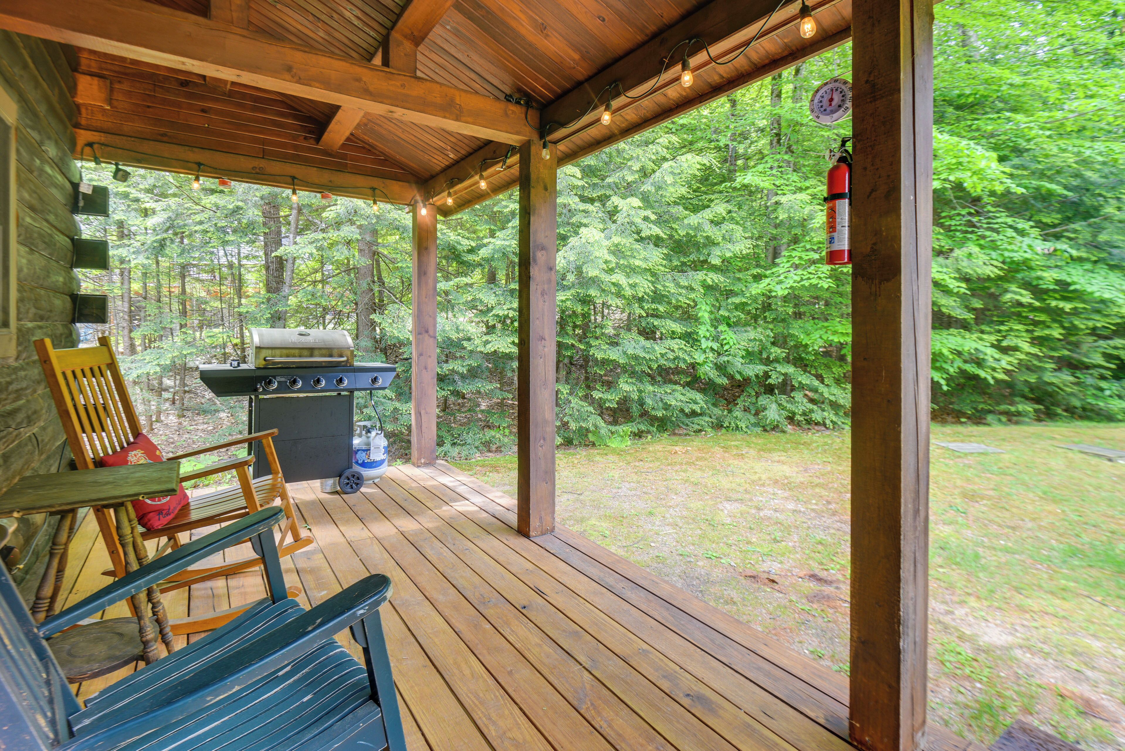 A covered porch with a grill and seating area, surrounded by a forest.