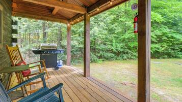 A covered porch with a grill and seating area, surrounded by a forest.