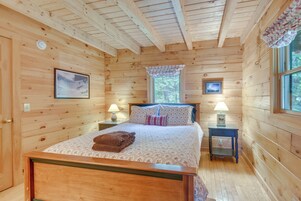 Un dormitorio de cabaña de madera con una cama, dos mesitas de noche con lámparas, una ventana con cortina y dos cuadros en las paredes.