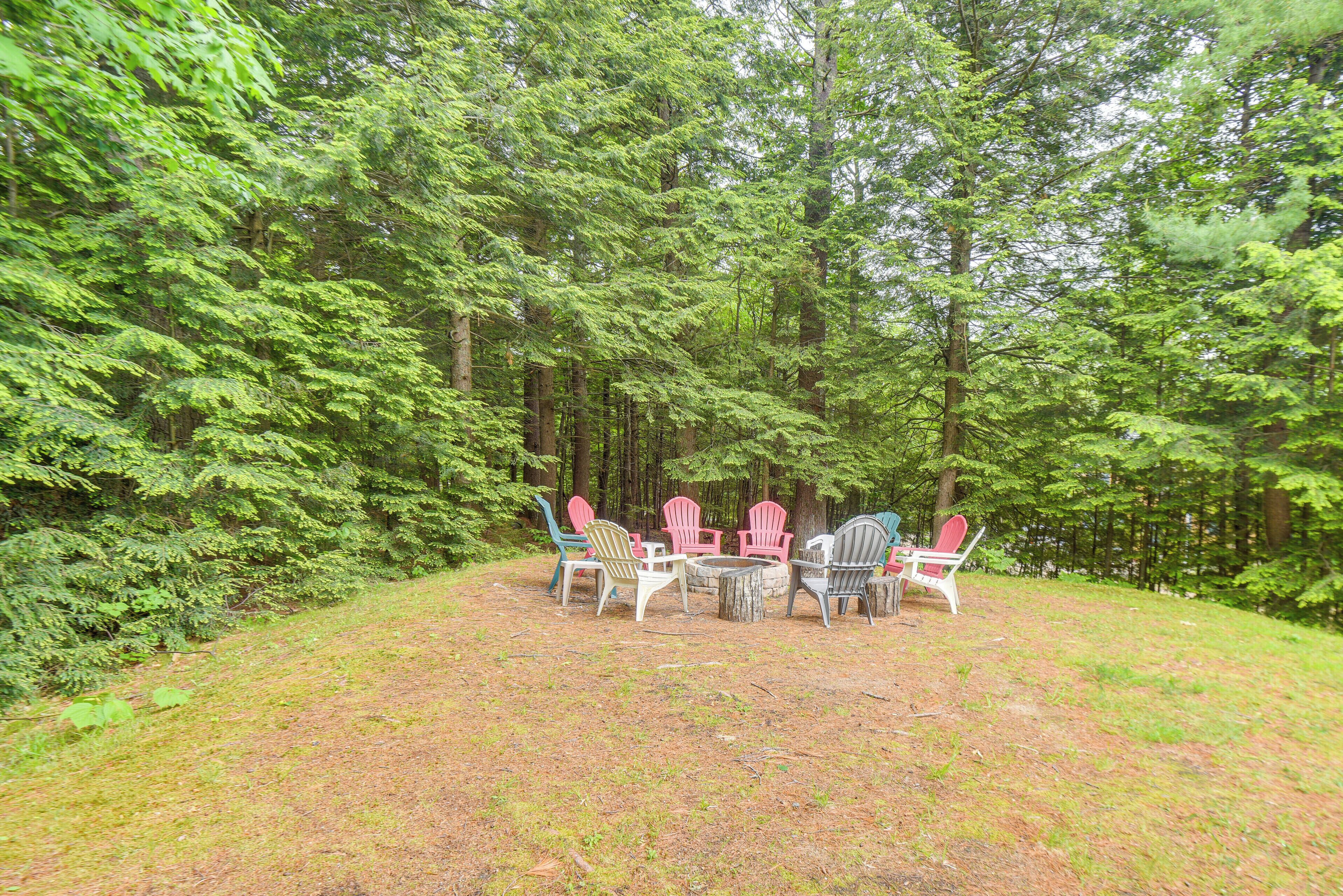 A group of Adirondack chairs arranged around a small fire pit in a forested area.