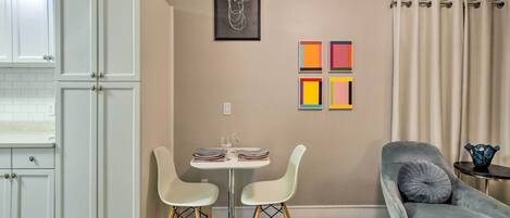 A dining area with a table set for two, a gray armchair, and a wall with a framed abstract art piece and colorful square paintings.