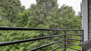 Un balcón con barandilla de metal, una silla de mimbre y vistas a árboles verdes y frondosos.
