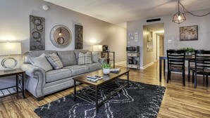 Una sala moderna con un sofá gris, una mesa de centro y un tapete negro. Hay un comedor con una mesa y sillas de madera.