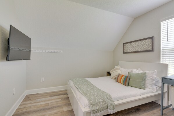 A bedroom with a white bed, a gray TV mounted on the wall, a small table, and a window with blinds. - Walk to Water: Family-friendly Surfside Beach Home (Freeport)