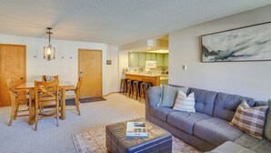 Ein Wohnzimmer mit grauem Sofa, einem Holzesstisch und Stühlen. Im Hintergrund ist eine Küche mit grünen Schränken und einer weißen Mikrowelle zu sehen.