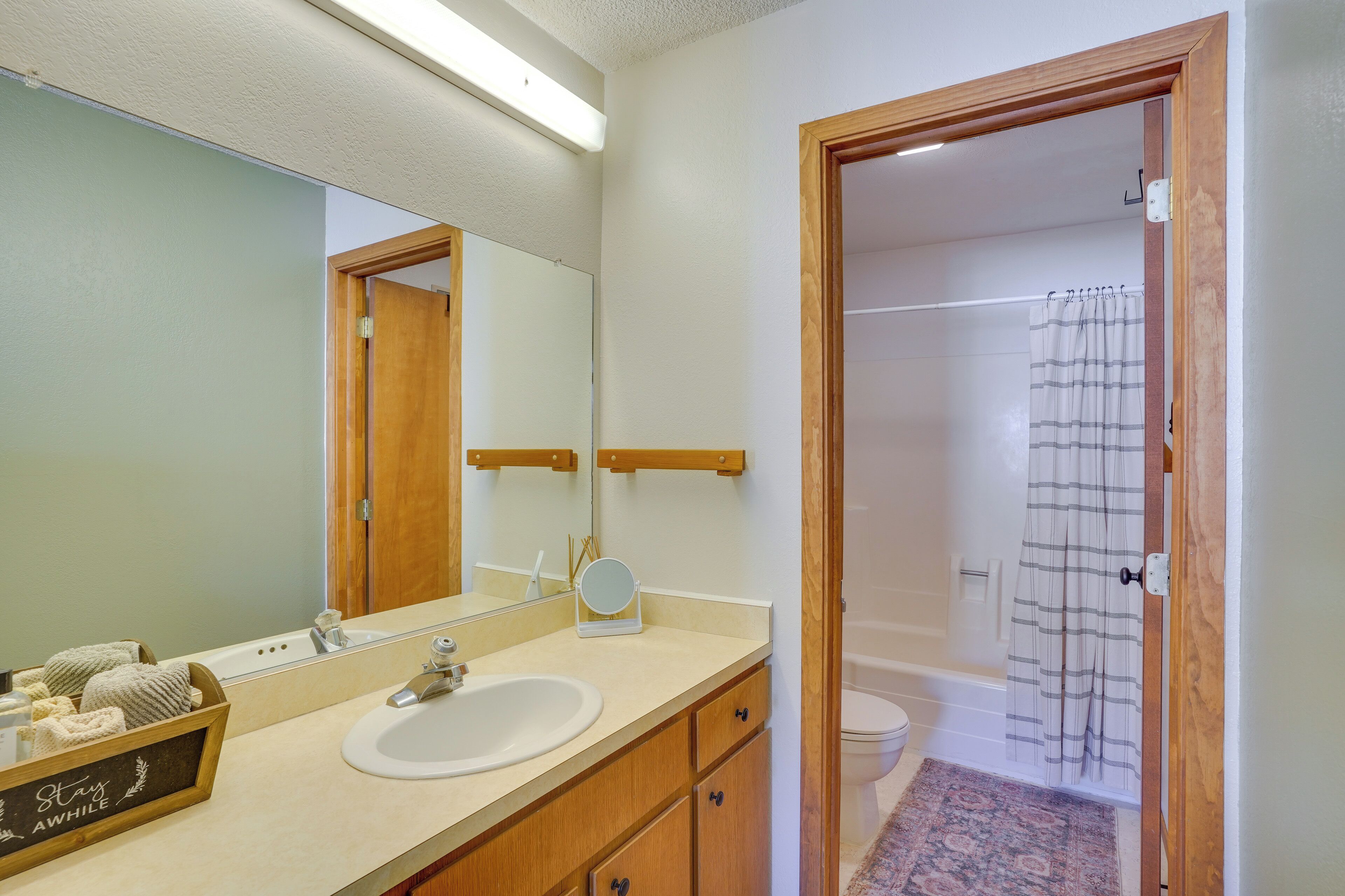 A bathroom with a sink, mirror, and wooden cabinets. There is a shower curtain and a rug in the adjacent bathroom.