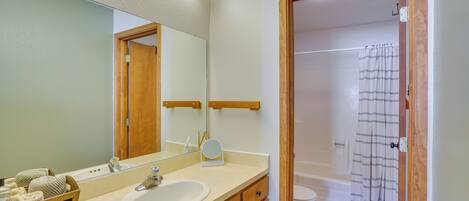 A bathroom with a sink, mirror, and wooden cabinets. There is a shower curtain and a rug in the adjacent bathroom.