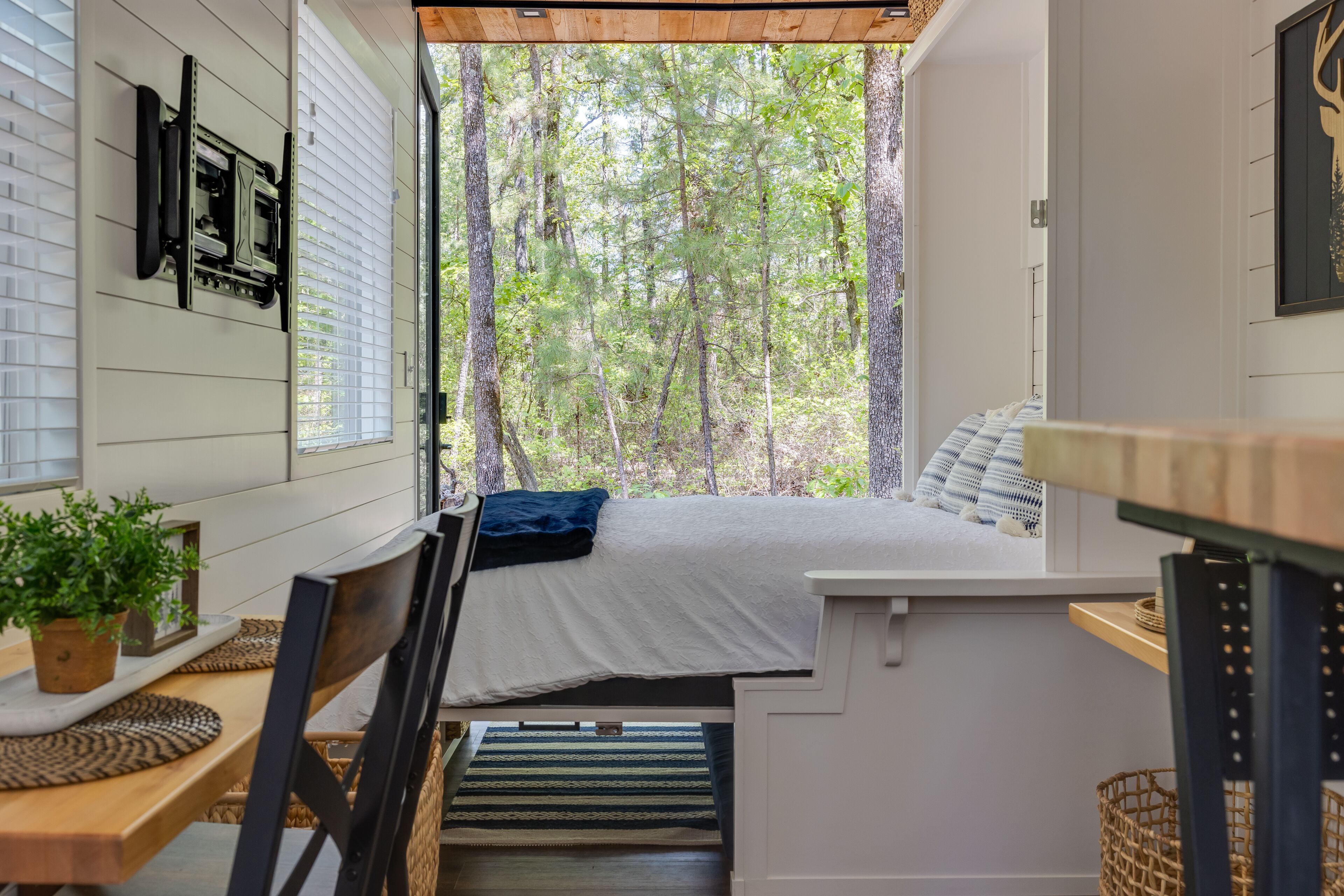 A compact bedroom with a bed, a small table, and a window overlooking a forest.