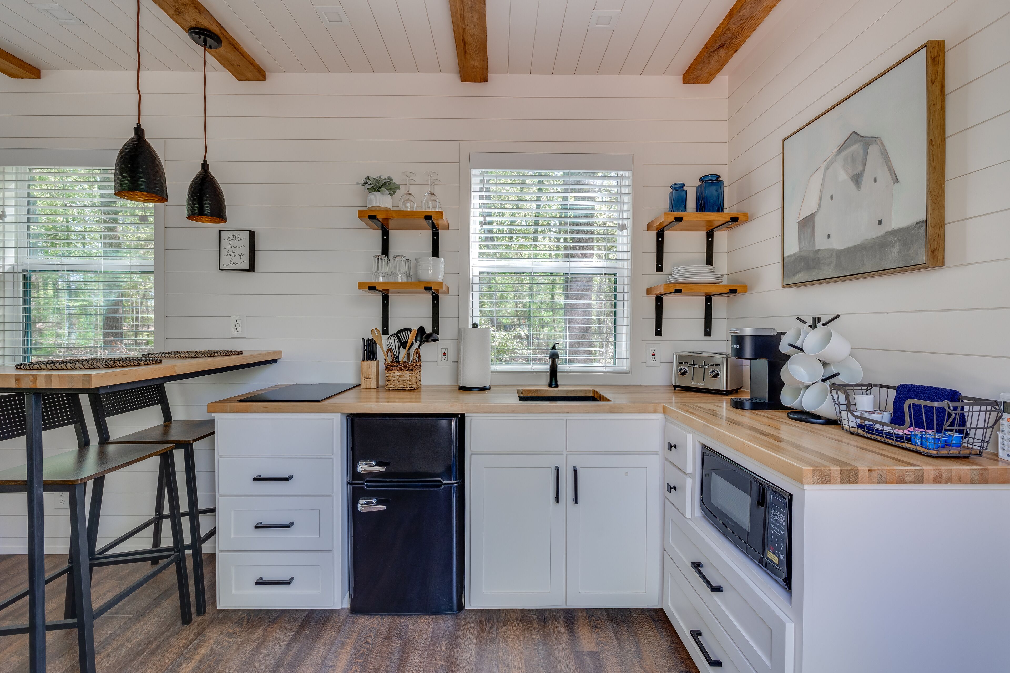 A modern kitchen with white cabinetry, a wooden countertop, and a black refrigerator. There are two pendant lights, a window with blinds, and a framed picture on the wall.