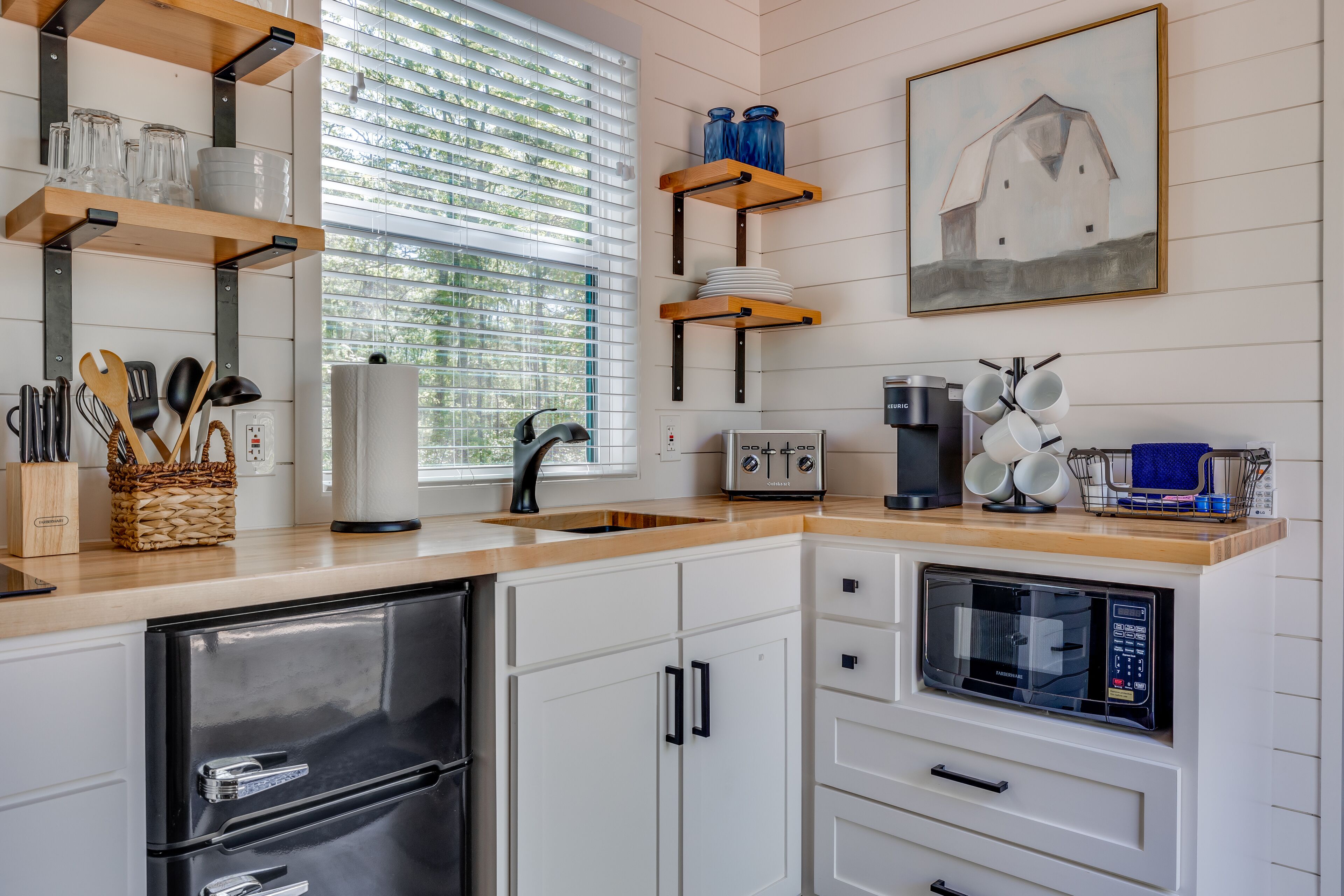 Cocina moderna con armarios blancos, un refrigerador de acero inoxidable, horno empotrado y microondas. Se observan estantes de madera con cristalería y un cuadro de un granero en la pared.