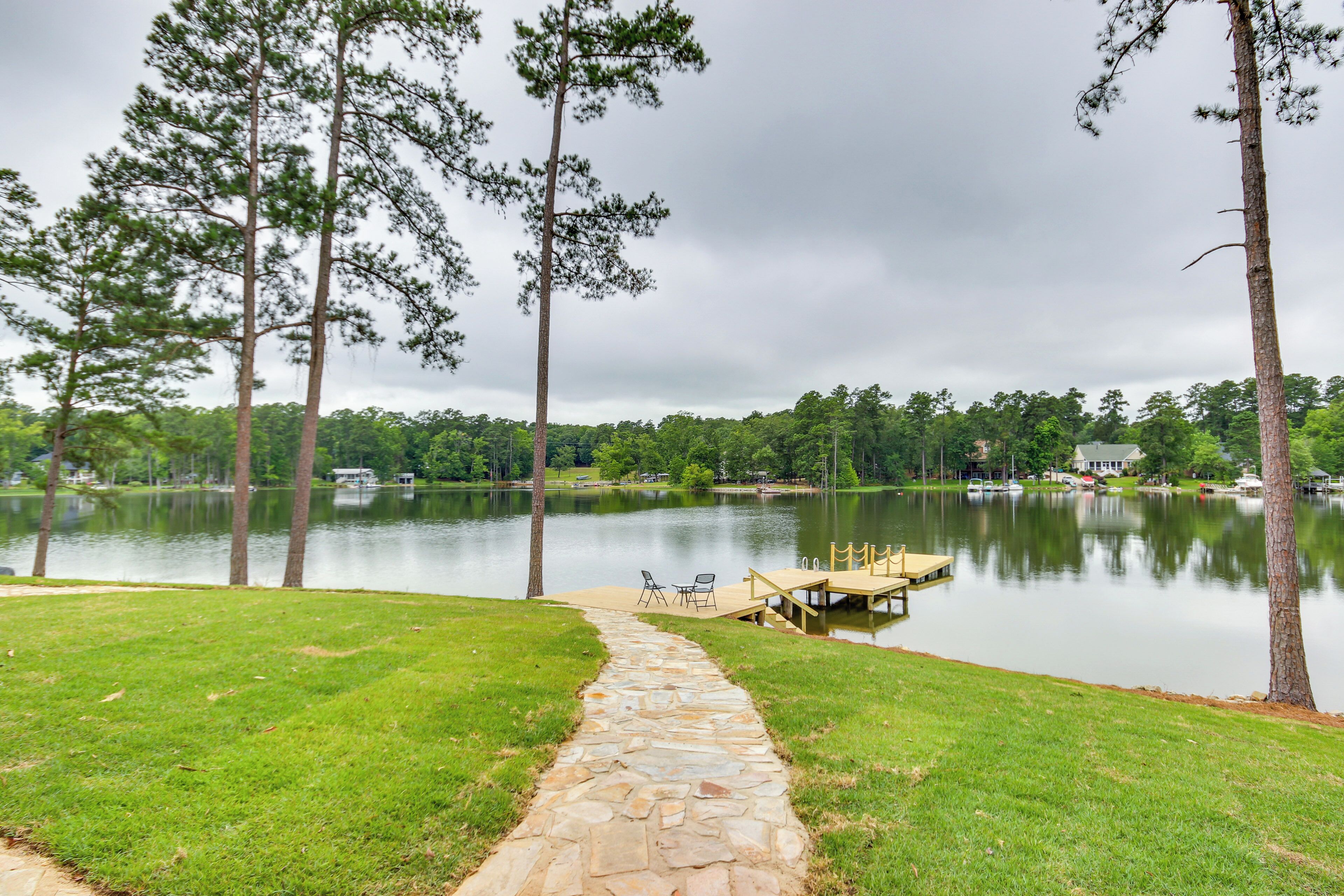 A stone pathway leading to a dock by a calm lake, surrounded by tall trees and a grassy area.