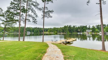 A stone pathway leading to a dock by a calm lake, surrounded by tall trees and a grassy area.