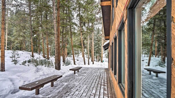 Una terraza de madera con bancos, rodeada de árboles altos y suelo cubierto de nieve.