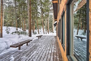 Una terraza de madera con bancos, rodeada de árboles altos y suelo cubierto de nieve.