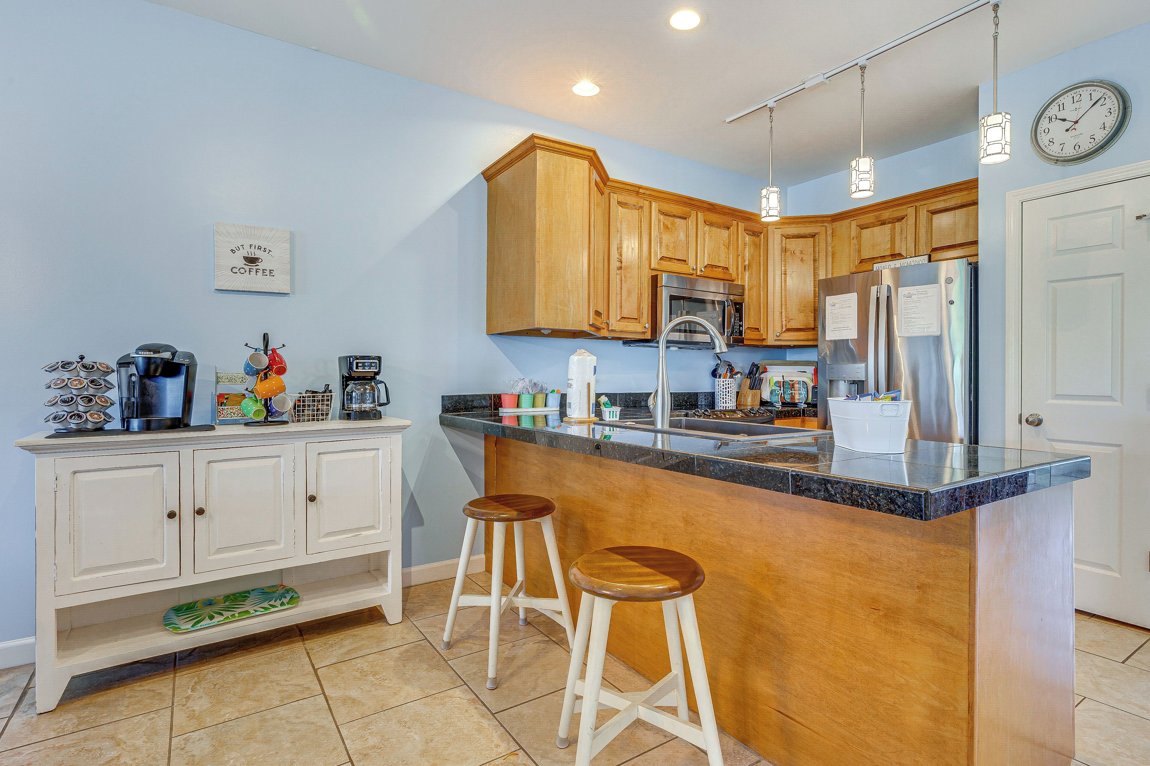 A kitchen with wooden cabinets, a stainless steel refrigerator, a bar counter, and two stools.