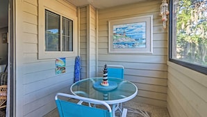 A small outdoor patio with a glass table and chairs, a framed painting of a lighthouse, and a decorative hanging ornament. - Family-friendly Condo w/ Pools + Tennis Courts! (Hilton Head Island)