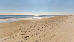 A wide sandy beach with footprints and tire tracks, leading to the ocean under a clear sky.