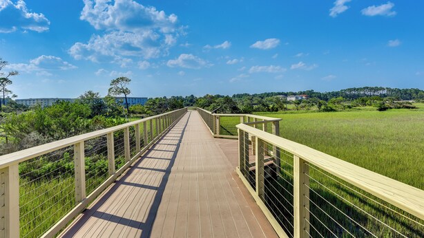A wooden boardwalk with metal railings, leading through a grassy field with trees and buildings in the background.