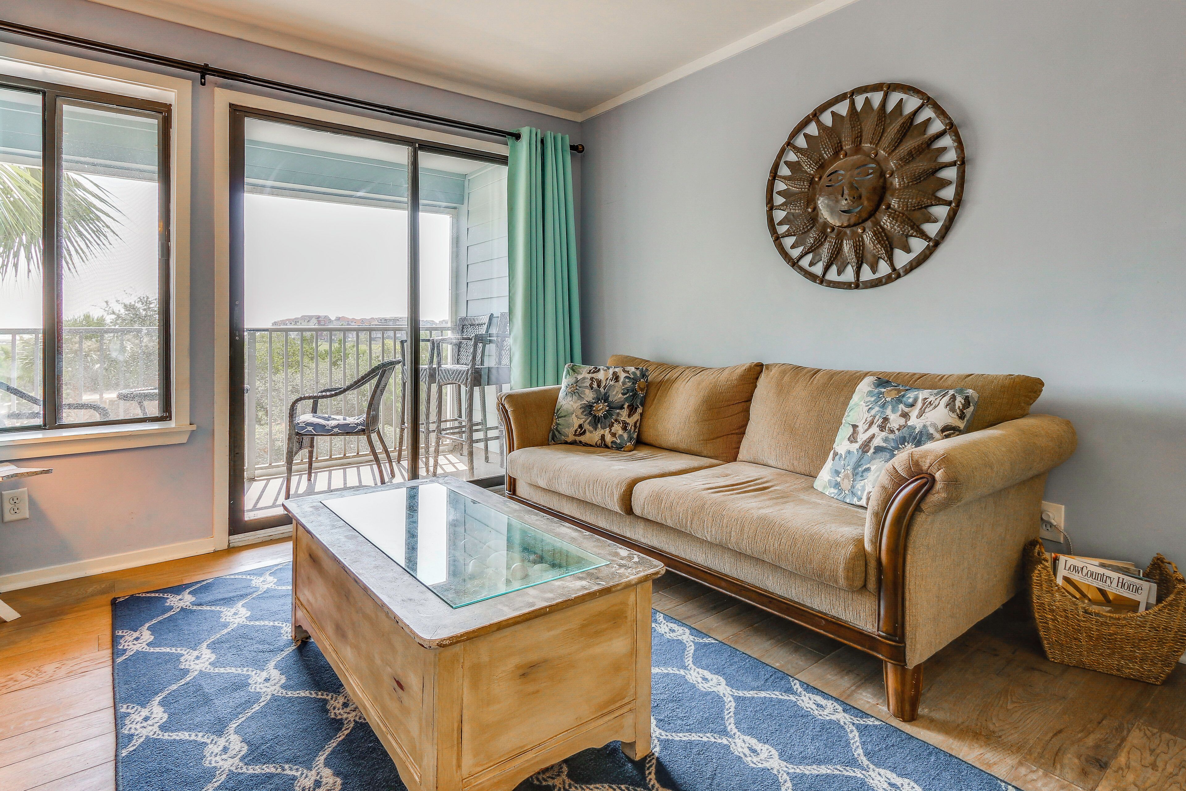 A living room with a sofa, a glass coffee table, a decorative wall piece, and a view through a sliding glass door to an outdoor area with chairs.