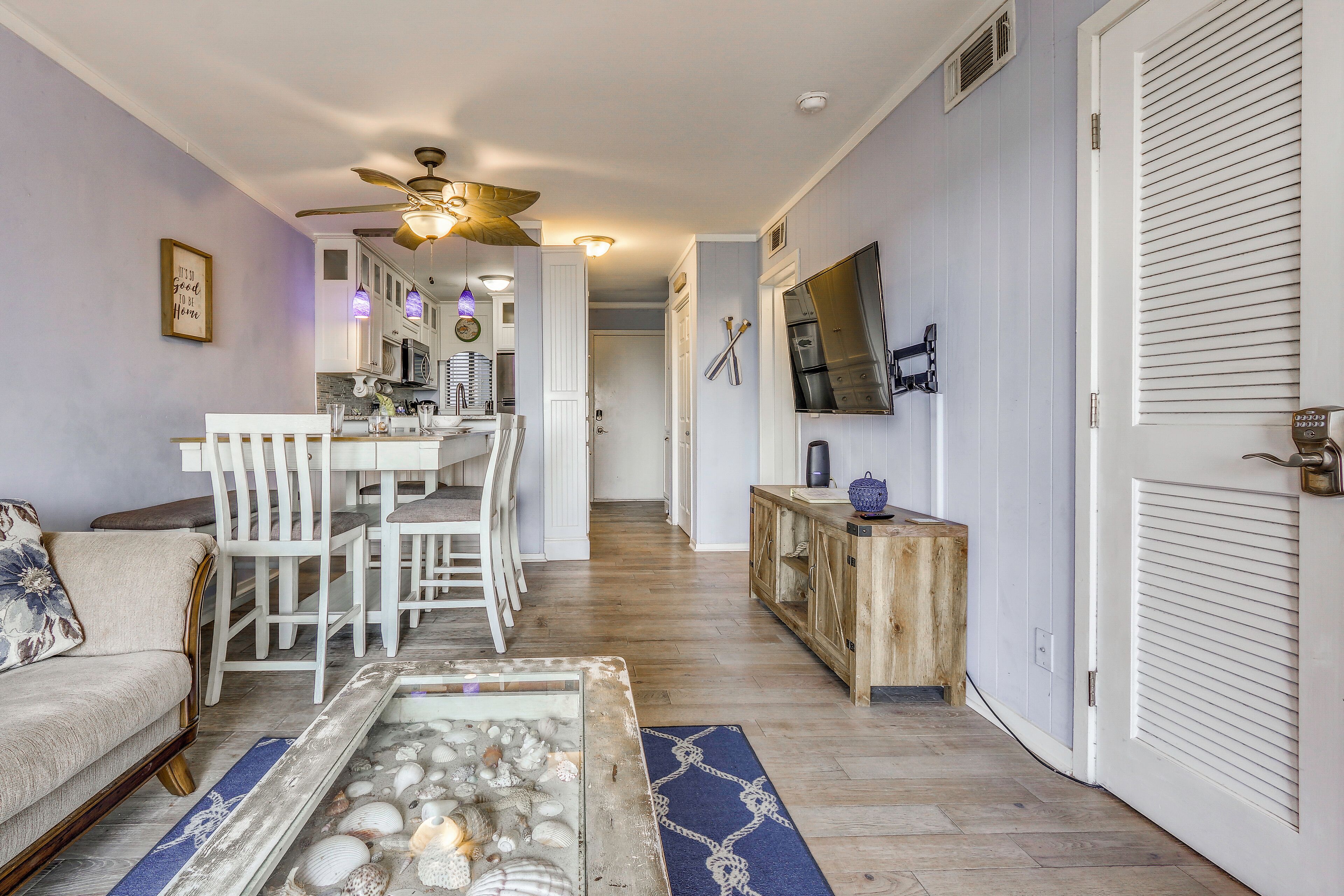 A living room with a sofa, a coffee table with seashells, a dining area with chairs, a kitchen with appliances, and a hallway leading to a bedroom.