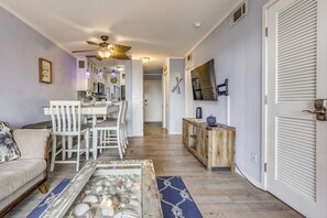 Uma sala de estar com sofá, mesa de centro com conchas, espaço de jantar com cadeiras, cozinha equipada e um corredor que dá para um quarto.
