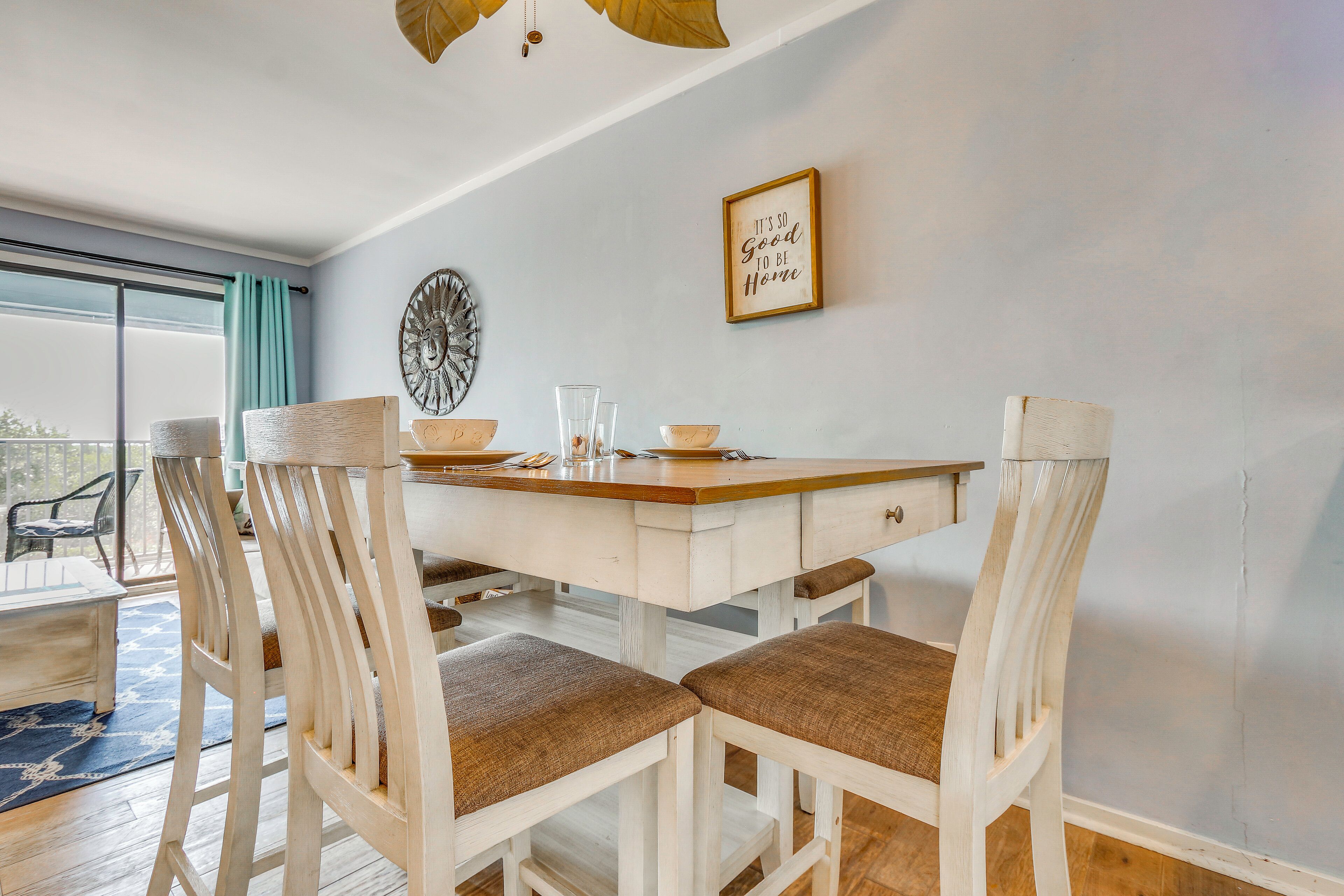 A dining area with a white table, four chairs with brown cushions, a decorative plate, and a framed sign on the wall.