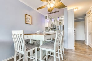 Un coin repas avec une table et des chaises en bois, un ventilateur de plafond et une cuisine en arrière-plan.