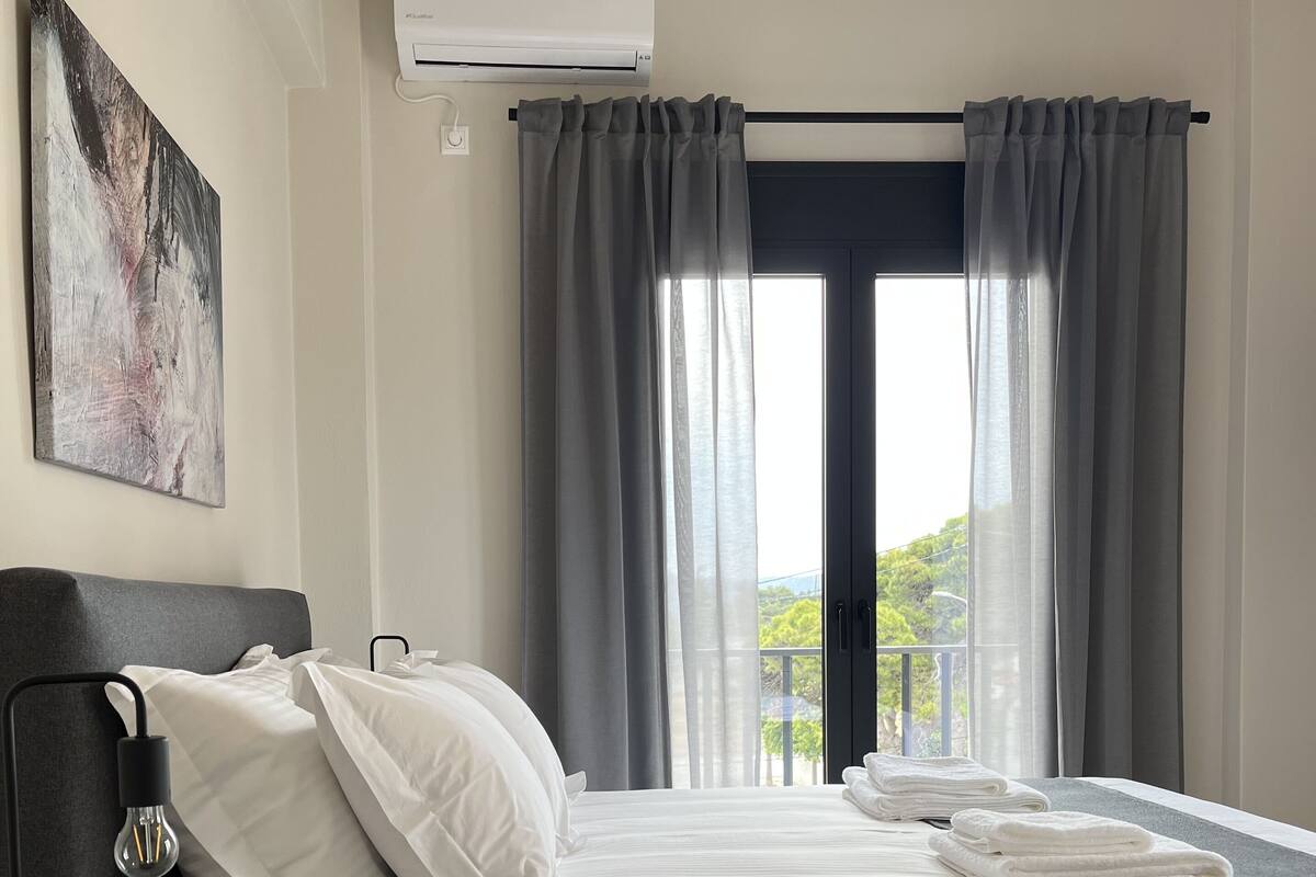 A modern hotel room with a bed, a bedside table with a lamp, an air conditioning unit, and a view of greenery through the window.