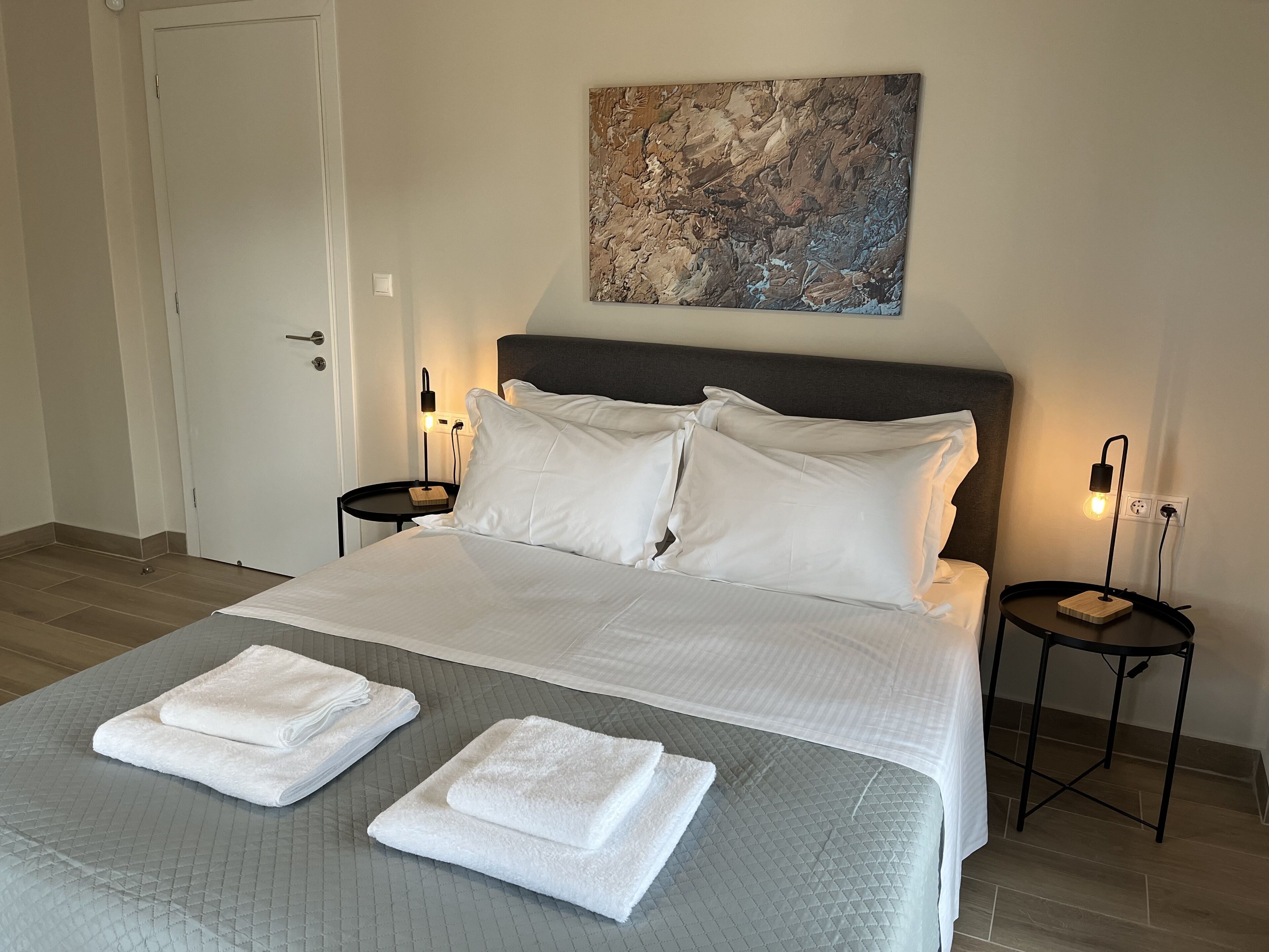 Ein ordentlich bezogenes Bett mit weißen Leinen und zwei gefalteten Handtüchern, eine graue gesteppte Bettdecke, ein moderner Kopfteil und ein abstraktes, strukturiertes Bild an der Wand darüber.