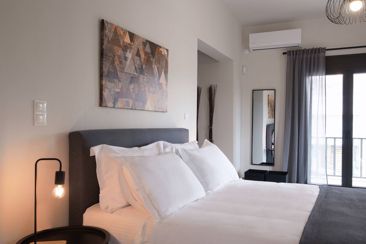 A modern hotel room with a large bed, a wall-mounted abstract painting, a bedside lamp, and a view of the outdoors through a window.