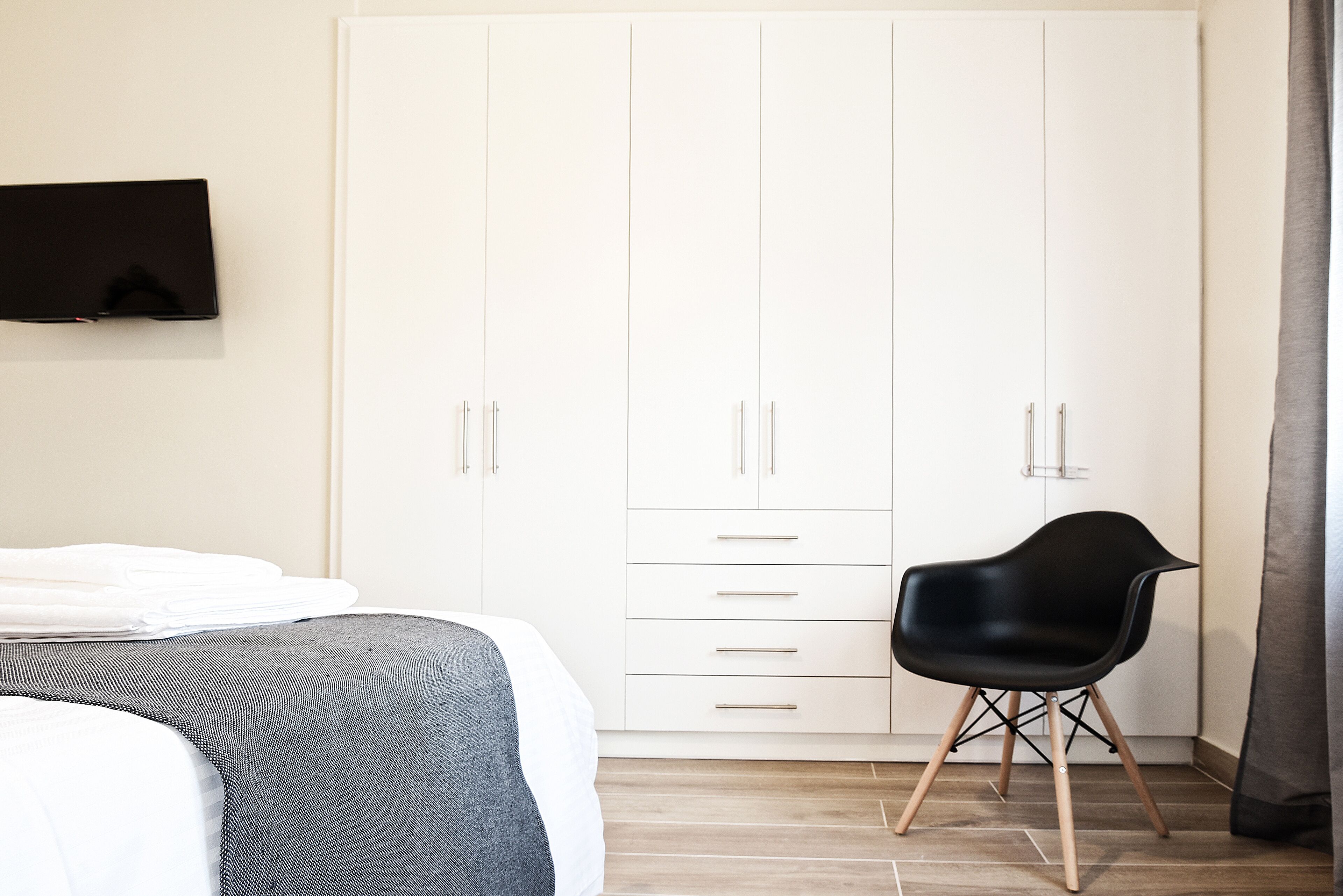 A bedroom with a bed, a TV mounted on the wall, a wardrobe with multiple drawers, and a modern chair.