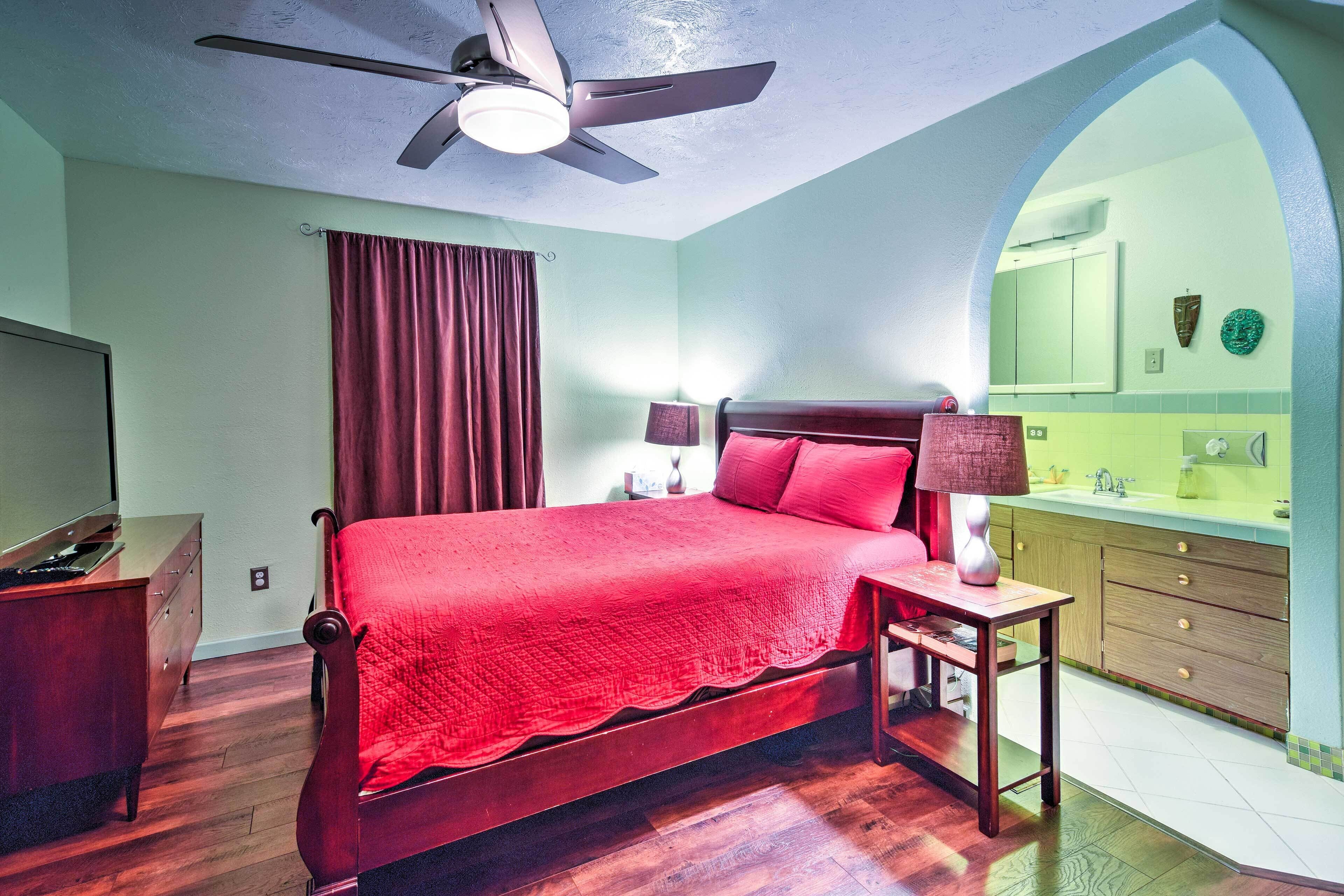 A bedroom with a wooden bed, a ceiling fan, a nightstand, a lamp, a dresser, and a bathroom with a sink and mirror.