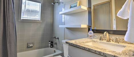 A modern bathroom with a bathtub, toilet, and sink. There are shelves above the sink and a window above the bathtub.