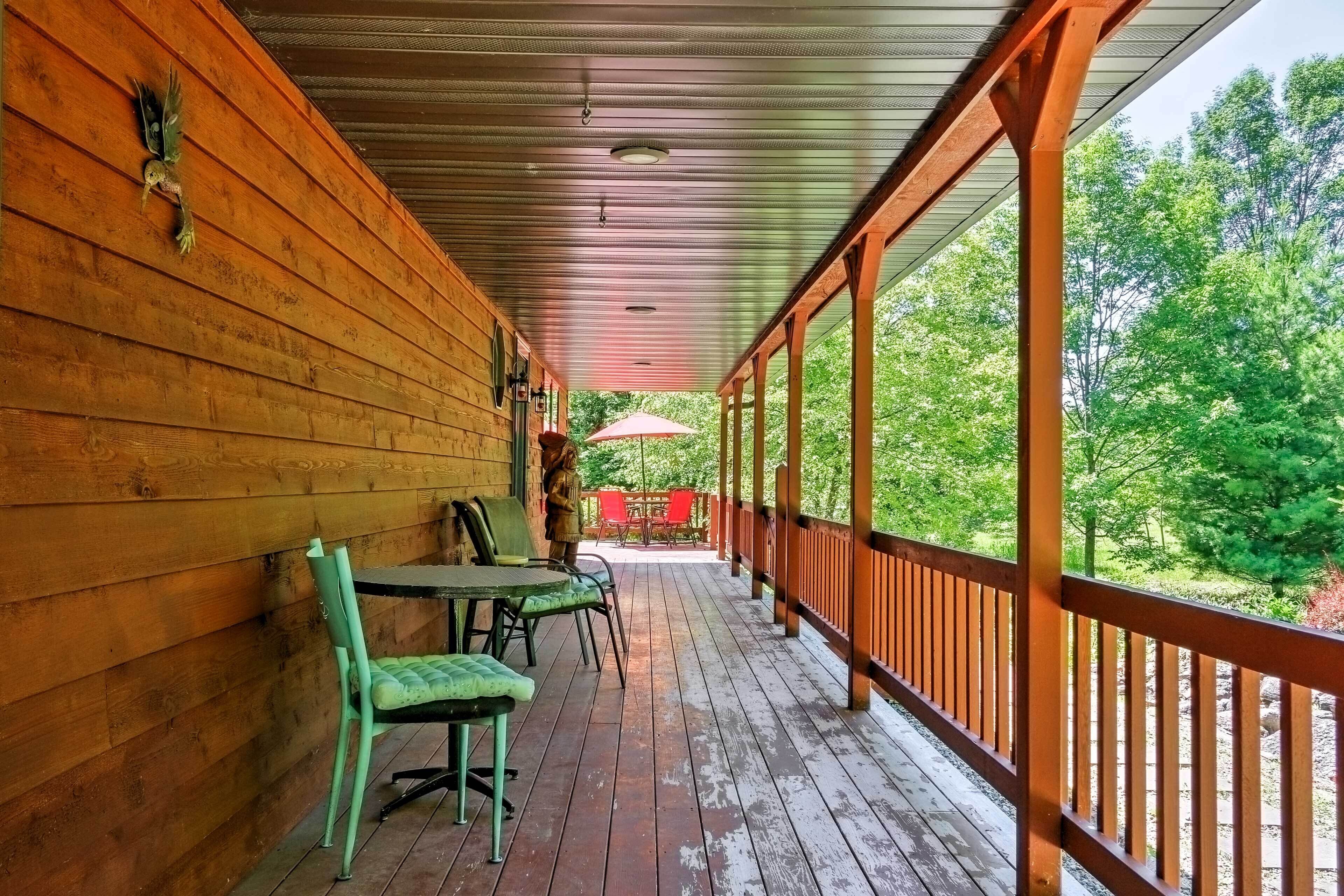 A covered porch with wooden walls and a railing, featuring a table and chairs, and a view of a wooded area.