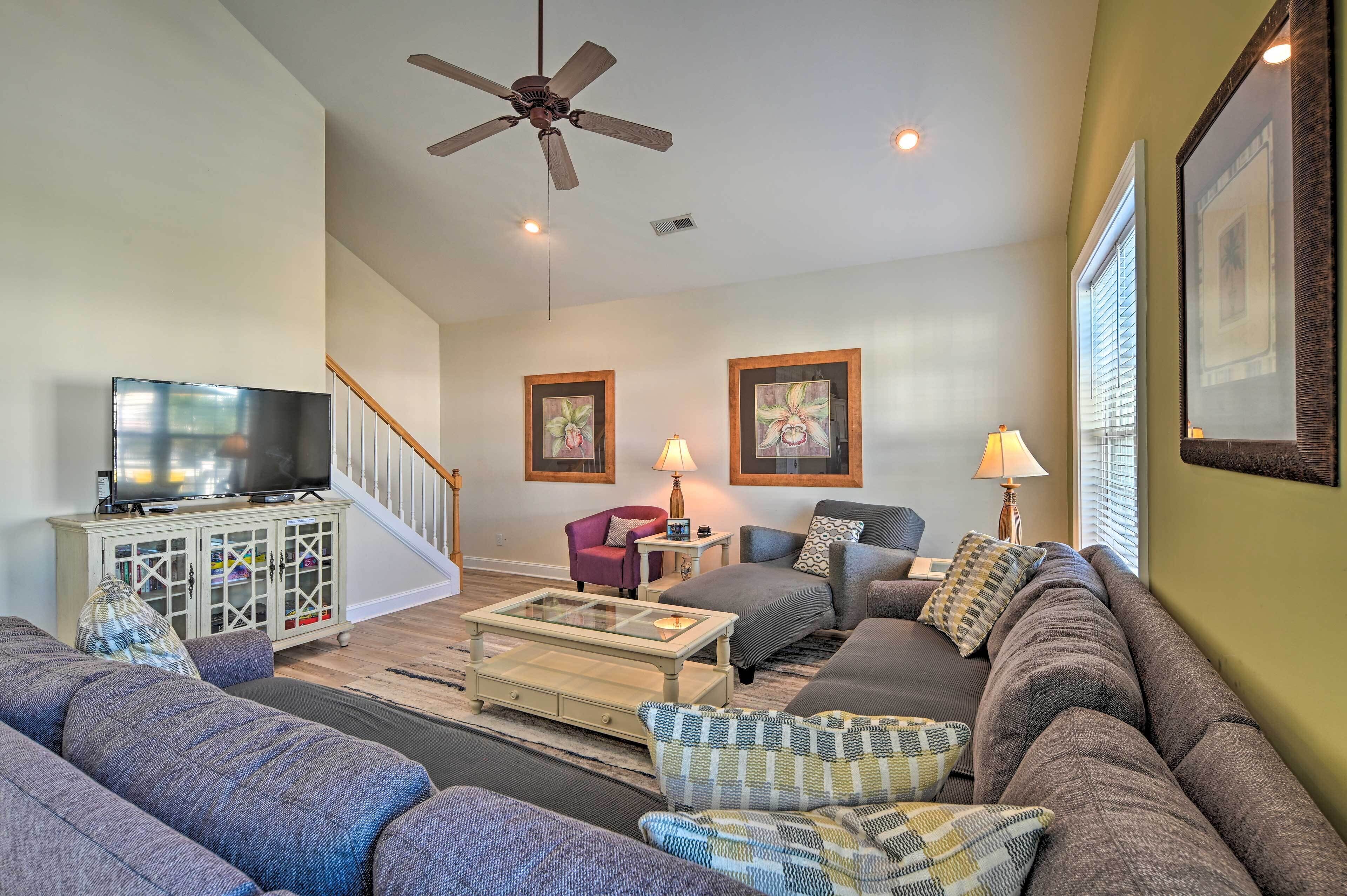 A spacious living room with a large sofa, a coffee table, and a television on a cabinet. There is a ceiling fan, a staircase, and framed artwork on the walls.