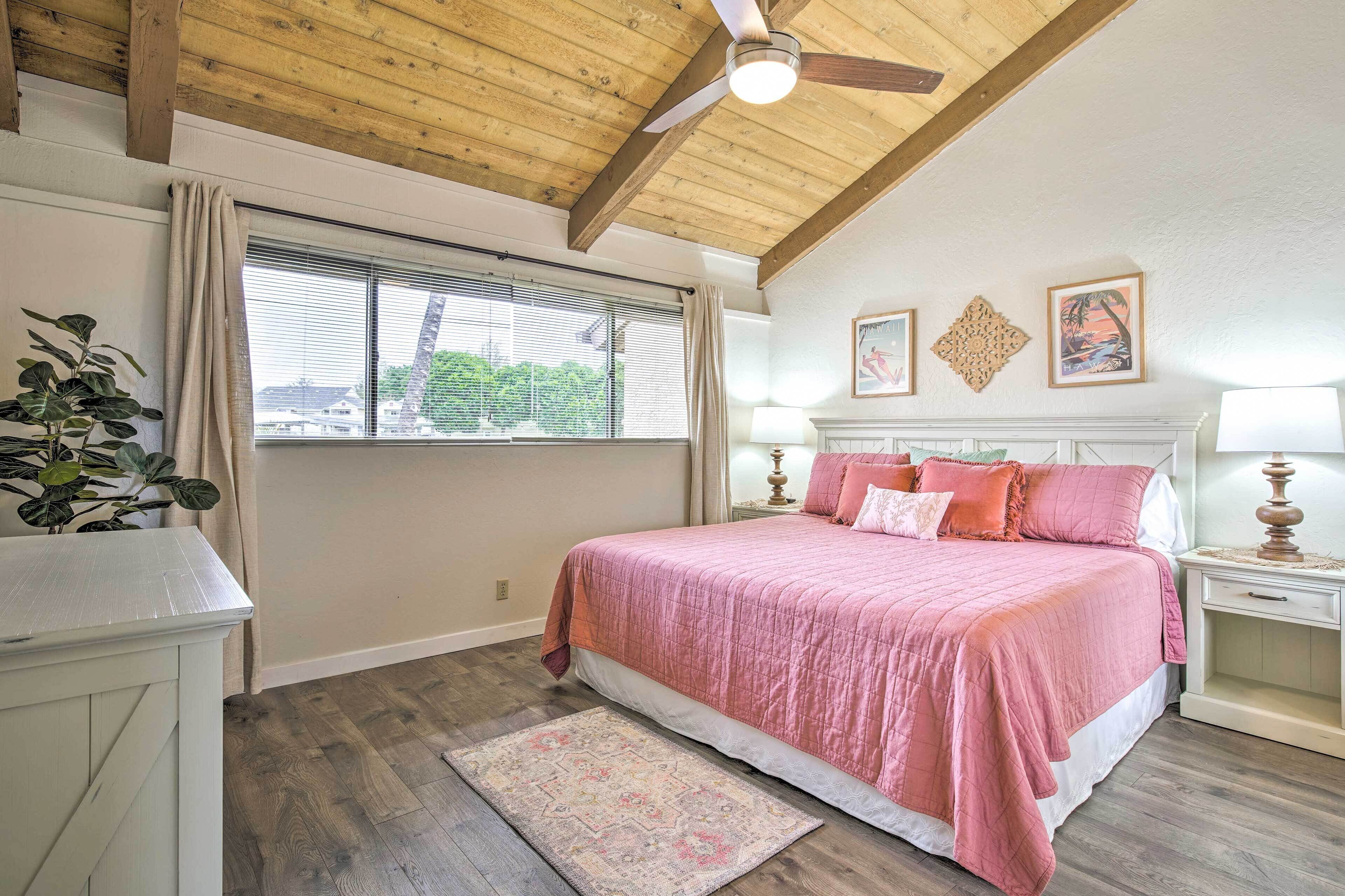 A bedroom with a wooden ceiling, a large bed with pink bedding, a nightstand with a lamp, and a window with curtains.