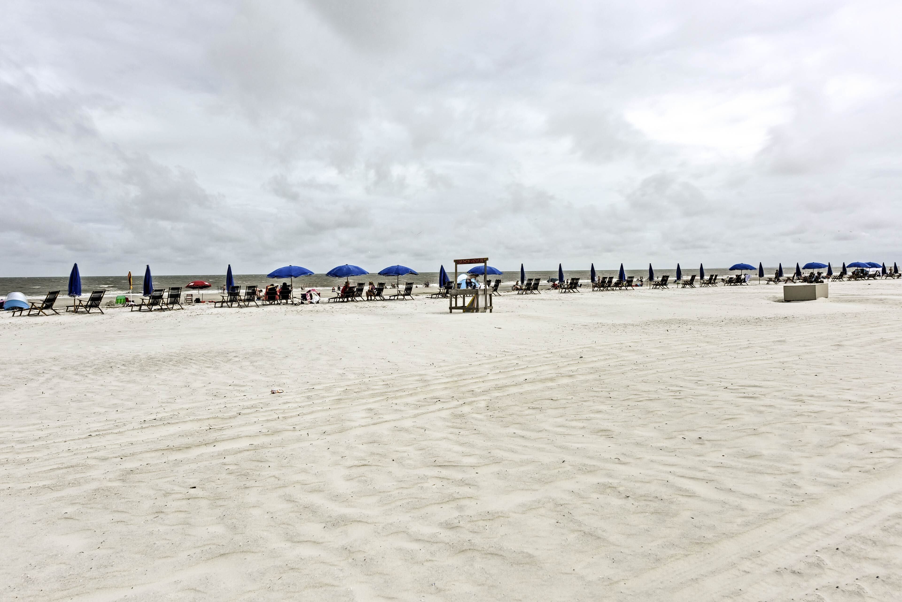 Ein Strand mit Reihen von Liegestühlen und Sonnenschirmen, einem Rettungsturm und dem Meer im Hintergrund.