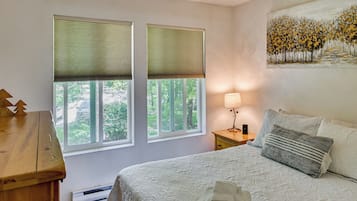 A bedroom with a bed, a nightstand, a lamp, and two windows with blinds.