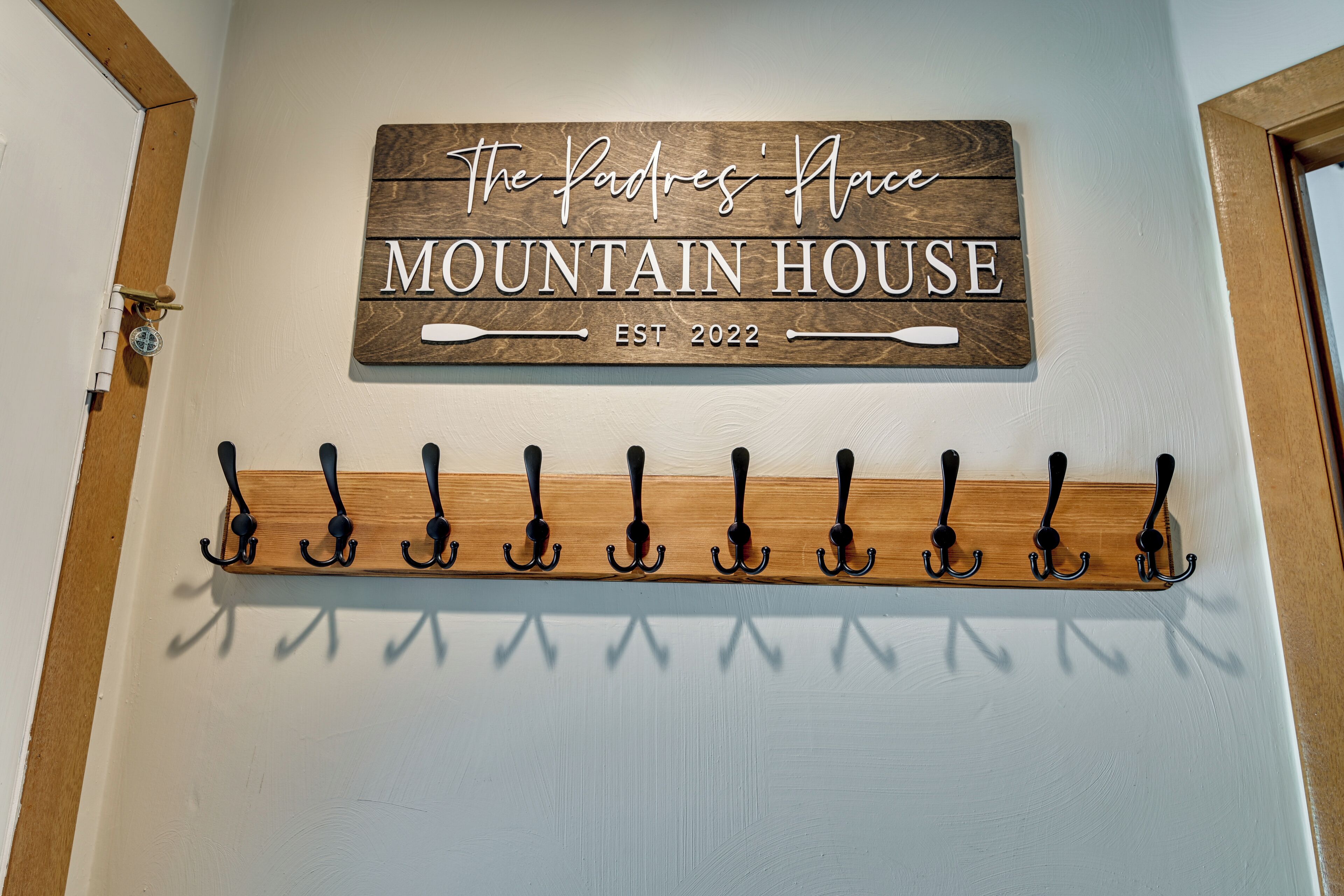 A wooden sign with 'The Padres' House Mountain House' and 'EST 2022' on a wall. Below is a black metal coat rack with hooks.