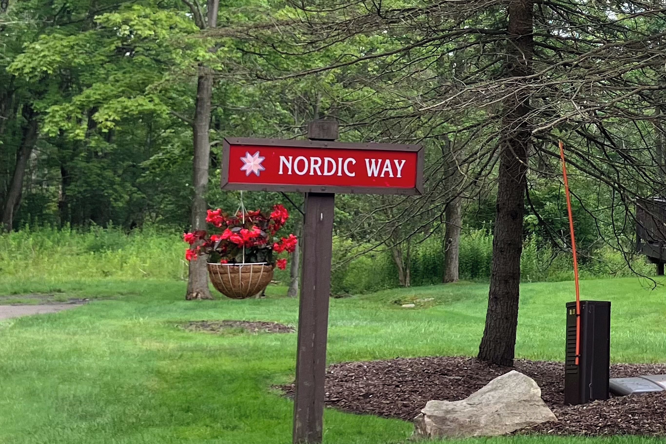 A sign for 'Nordic Way' with a red background and white lettering, a hanging flower basket, and a stone pathway.