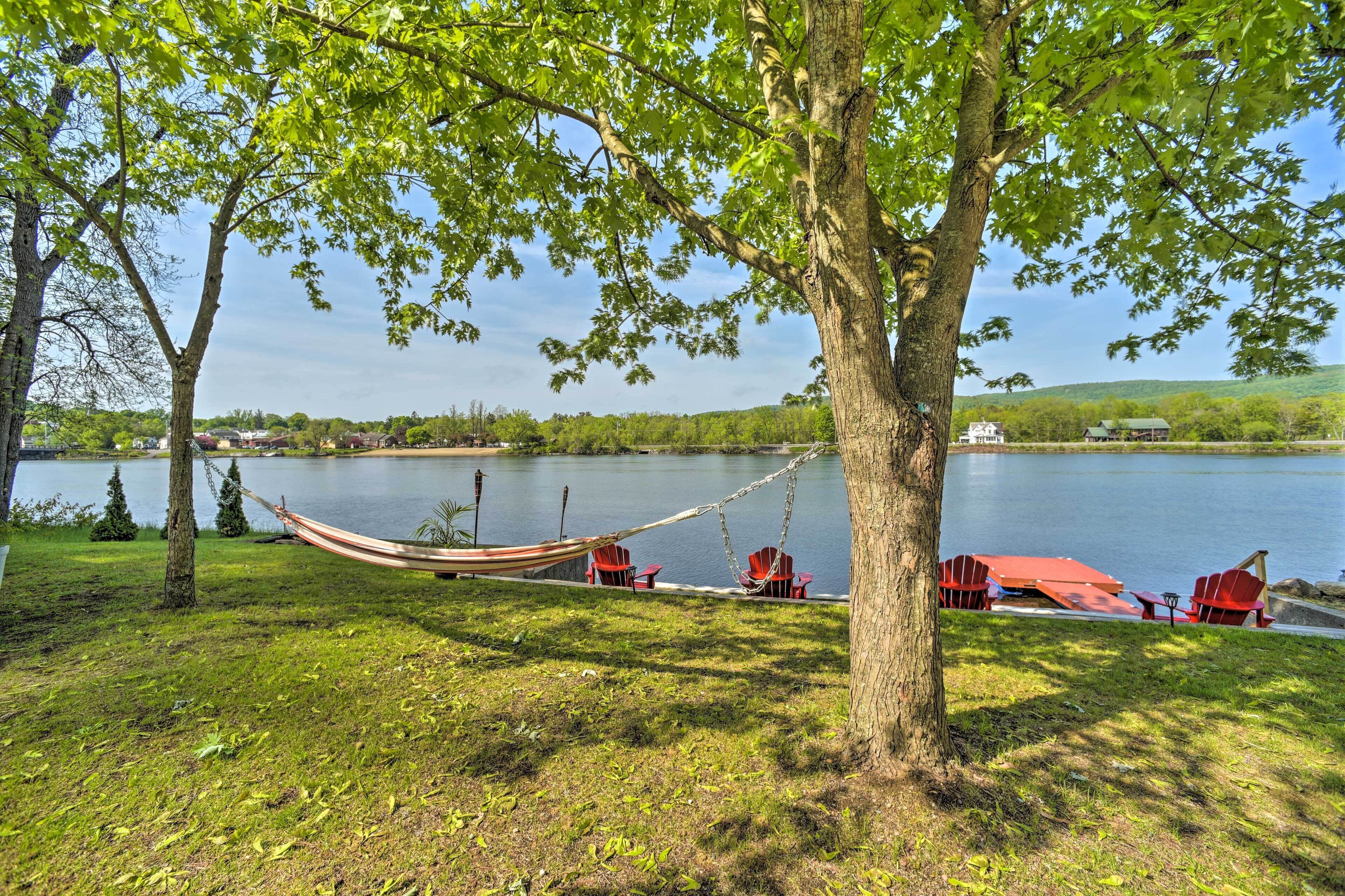Una hamaca bajo un árbol junto a un lago, con sillas rojas y un muelle.