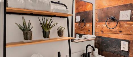 Un baño moderno con pared de madera, un lavamanos blanco, un espejo y estantes con plantas y artículos de aseo.