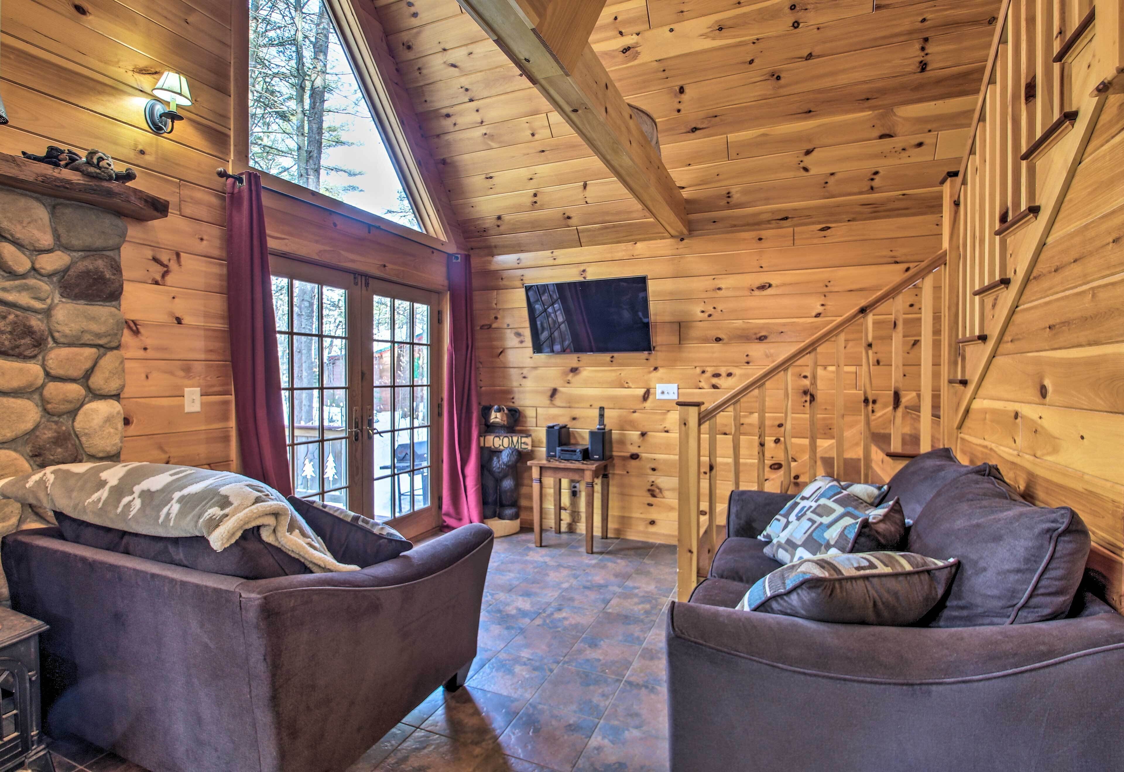 A cozy cabin interior with two sofas, a stone fireplace, a TV, and a staircase leading to a loft area.