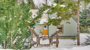 Eine Holzhütte mit einer Holztreppe, die zu einer gelben Tür führt, umgeben von immergrünen Bäumen und Schnee.