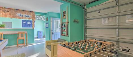 A room with a foosball table, a bar area labeled 'Tiki Bar', and a garage door with a sign indicating it is not to be opened manually.