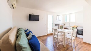 A modern living room with a sofa, a dining table with chairs, a kitchen area with a stove, and a flat-screen TV mounted on the wall. - Viewsuvio Terrace at historical center (Napoli)
