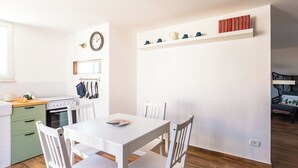 A compact kitchen with white cabinetry, a dining table, and chairs. There is a clock on the wall and a shelf with books and cups. - Viewsuvio Terrace at historical center (Napoli)