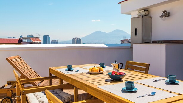 A wooden table with a view of a mountain, a bowl of fruit, and blue cups on a balcony. - Viewsuvio Terrace at historical center (Napoli)