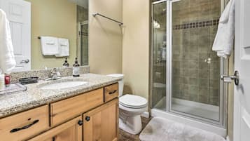 A bathroom with a granite countertop sink, wooden cabinets, a toilet, and a glass-enclosed shower.