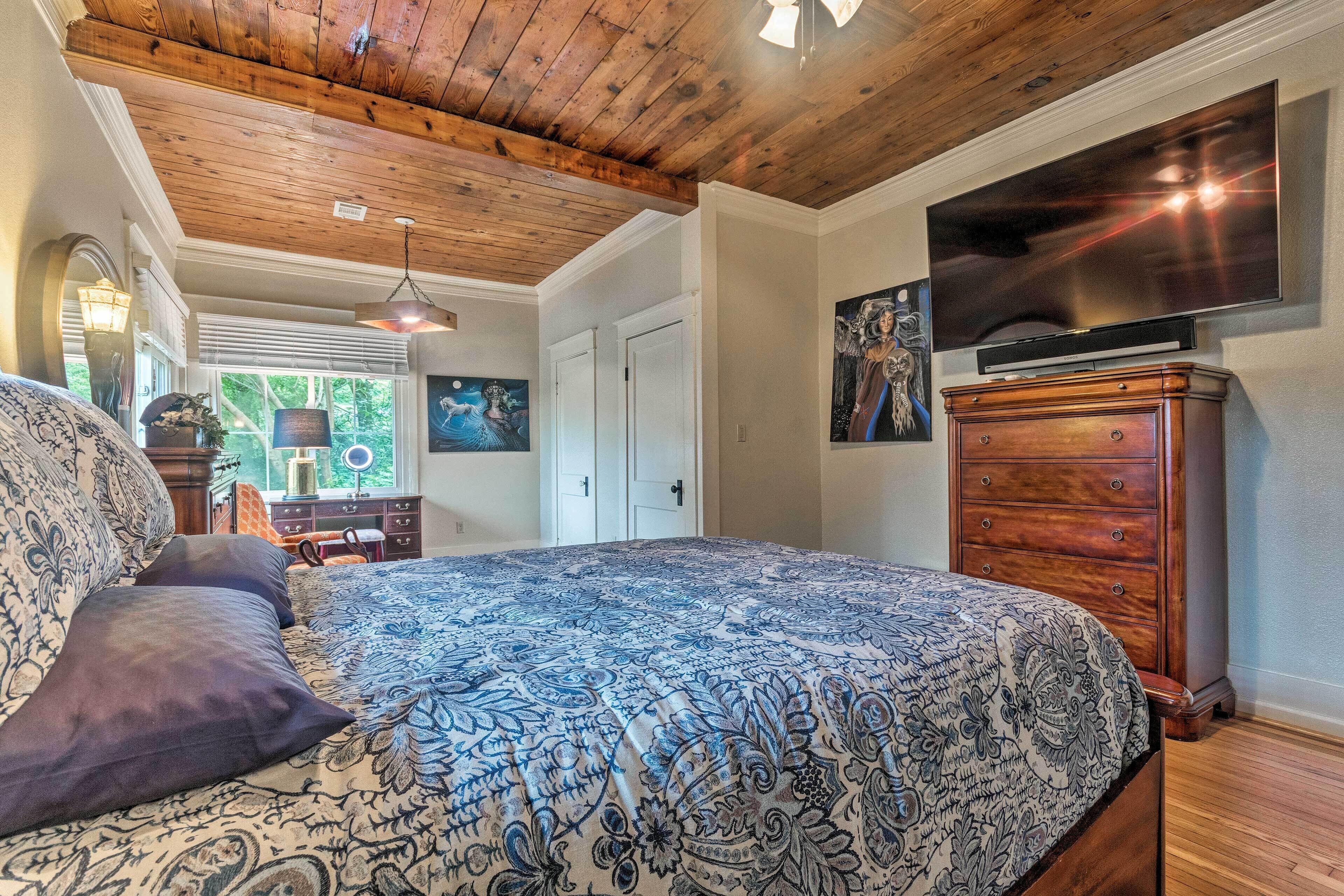 A bedroom with a large bed, wooden ceiling, a dresser, a television, and artwork on the walls.